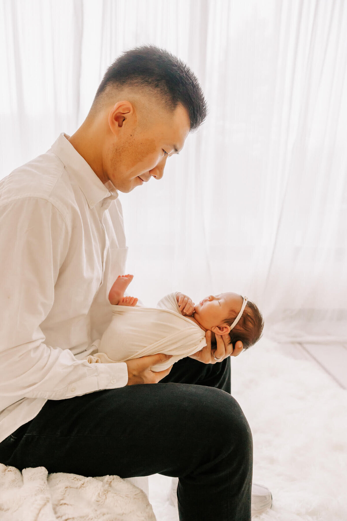 portrait of a dad wearing a white shirt and black pants sitting on the edge of a bed. he is looking down at his newborn baby girl