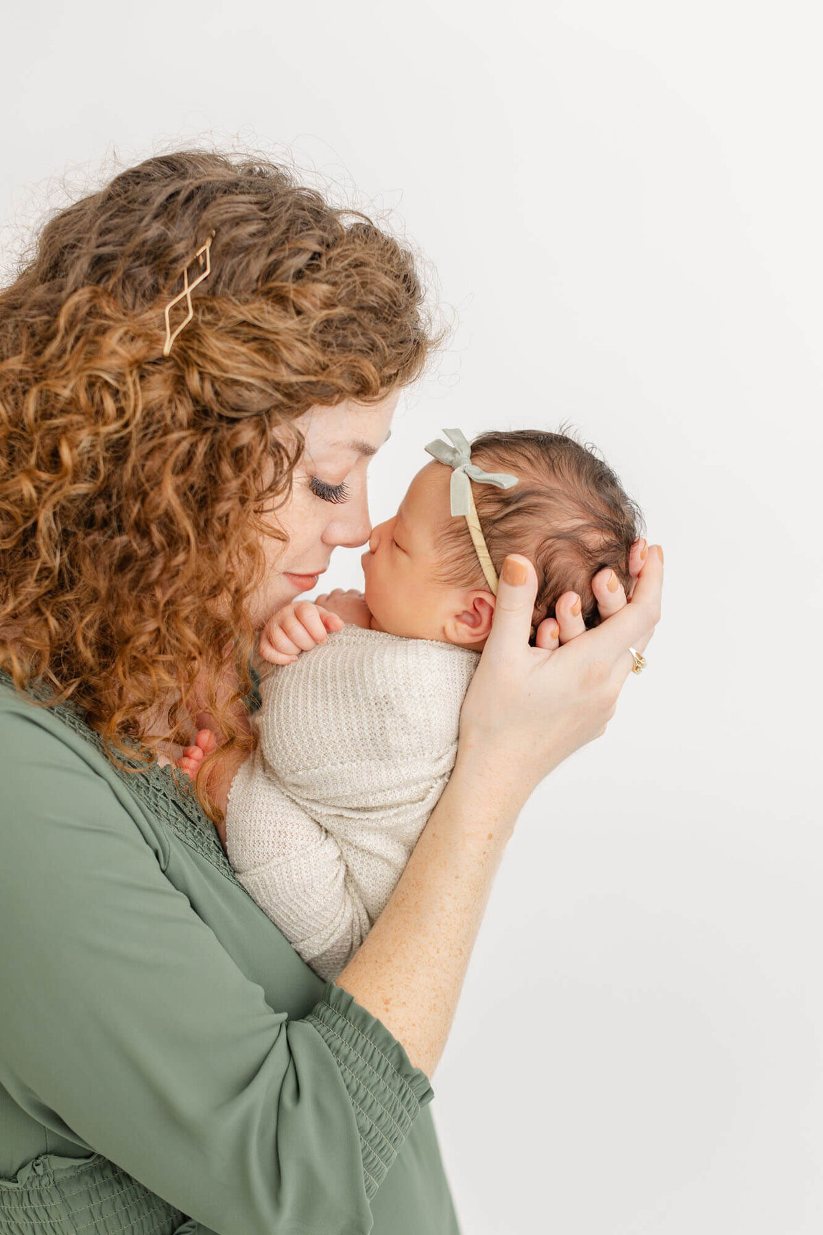 Mom is nose to nose with her sweet newborn baby girl. Mom is wearing a gorgeous green baltic born dress and baby is wrapped in beige with a small green velvet bow on her head