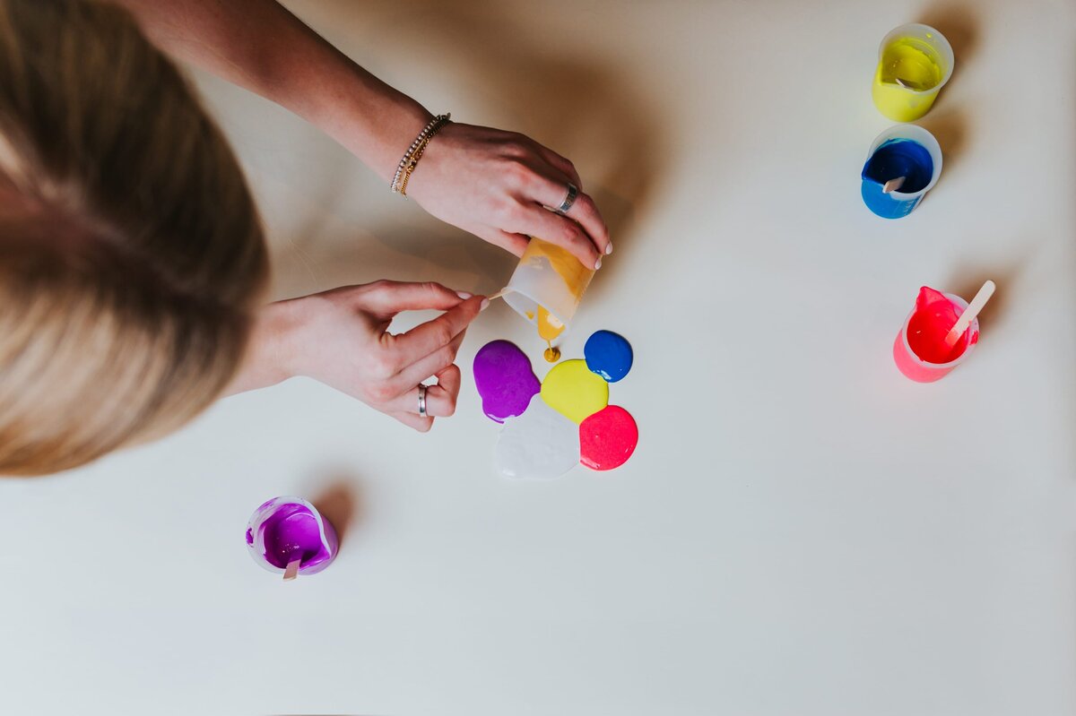 Overhead view of woman pouring puddles of vibrant paint onto white surface.