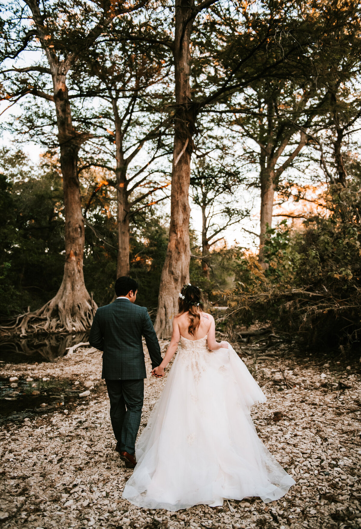 Couples Photography, Man in a navy blue suit holding hands with a woman in a wedding dress as they walk through a rocky creek with large Cypress trees.