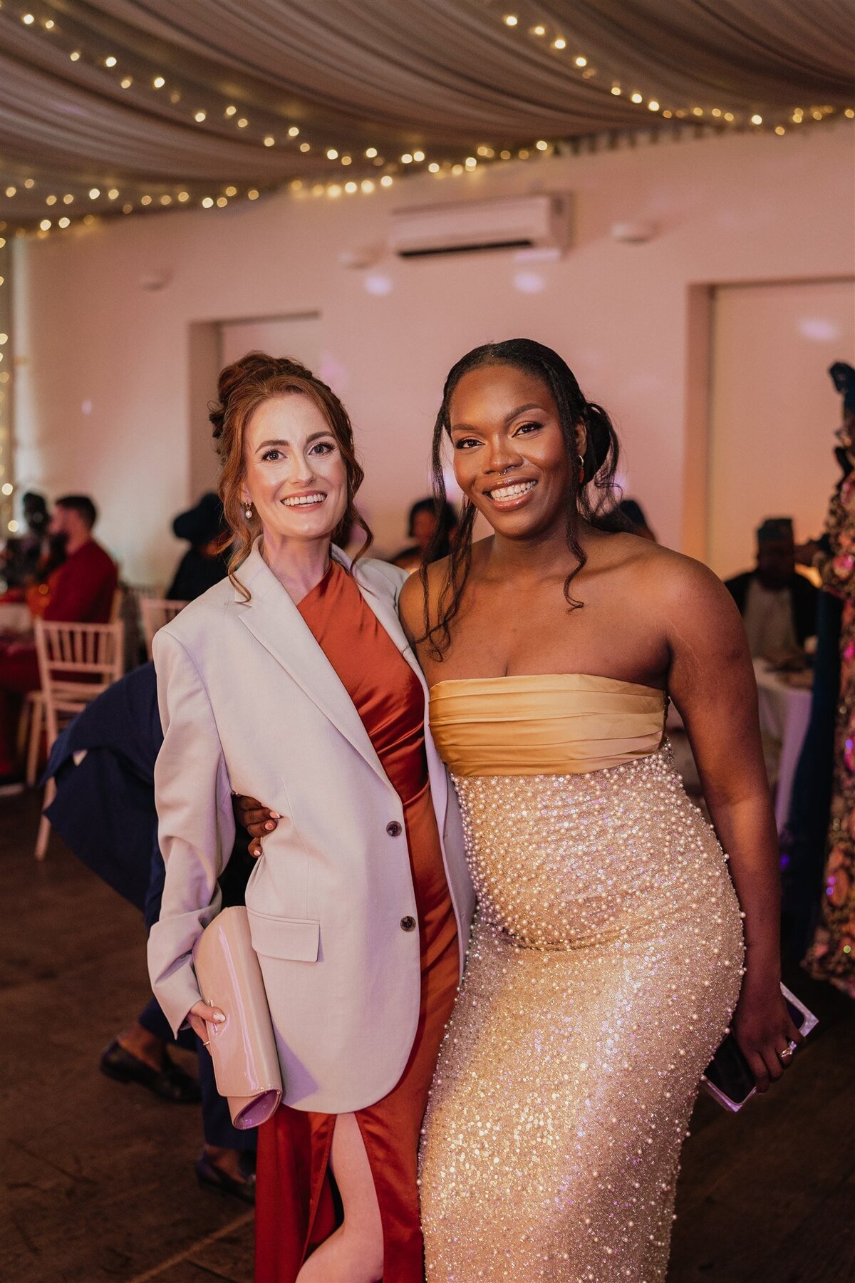 Two women stand side by side and pose for a photograph. The woman on the left wears a dress and an oversized blazer.