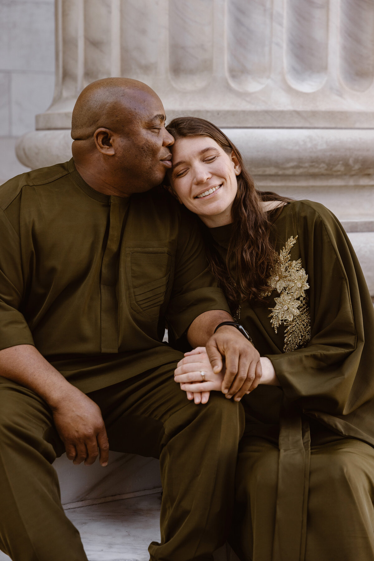 A man kisses his partner’s forehead while they sit together in front of marble columns, holding hands and smiling softly.