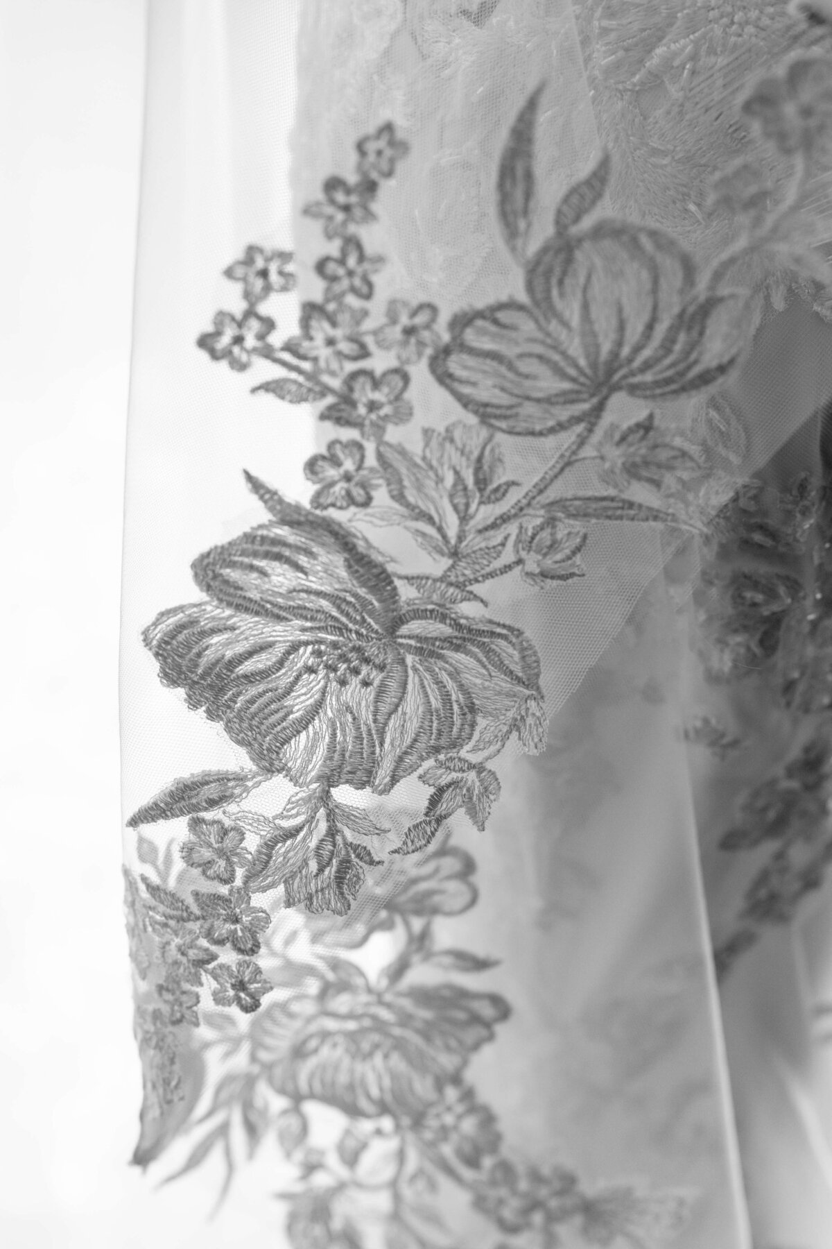 Close-up of a delicate lace fabric featuring intricate floral embroidery in a grayscale color scheme. The design highlights large blossoms and leaves, creating an elegant and detailed pattern.