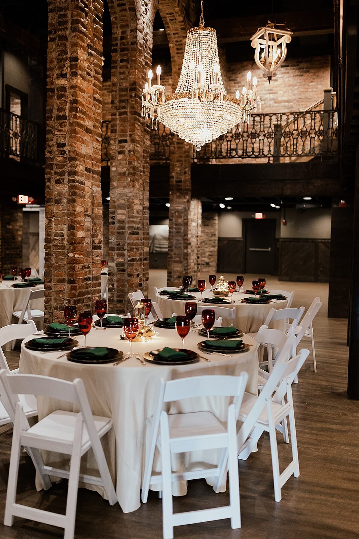 White garden chairs surround tables with white linens. The tables are set with a black charger topped with an emerald green napkin. On either side of the plate is gold flatware and a red water goblet. The venue has dark hardwood floors, exposed brick arches and and assortment of crystal chandeliers.