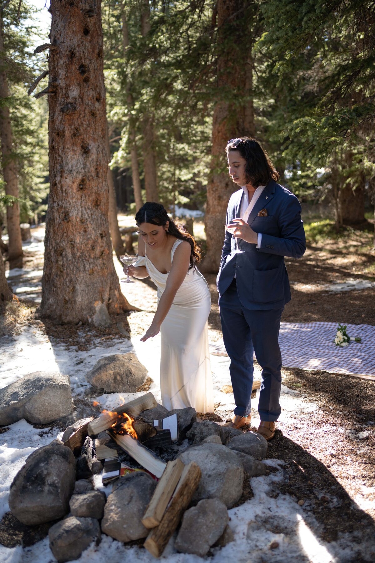 A newlywed couple enjoys an intimate picnic in a forest setting, sitting on a checkered blanket beneath tall trees. The bride, wearing a white dress, a beige coat, and hiking boots, smiles as she cuts a small white wedding cake with delicate floral decorations. Her dark hair is styled in loose waves. The groom, dressed in a navy blue suit with an unbuttoned white shirt, leans back comfortably, watching her with a warm expression. His outfit is accented with playful socks featuring a red and blue design. A bouquet of white and yellow flowers rests beside them on the blanket. Sunlight filters through the trees, casting a soft, natural glow on the moment, blending elegance with adventure in a serene wilderness setting.