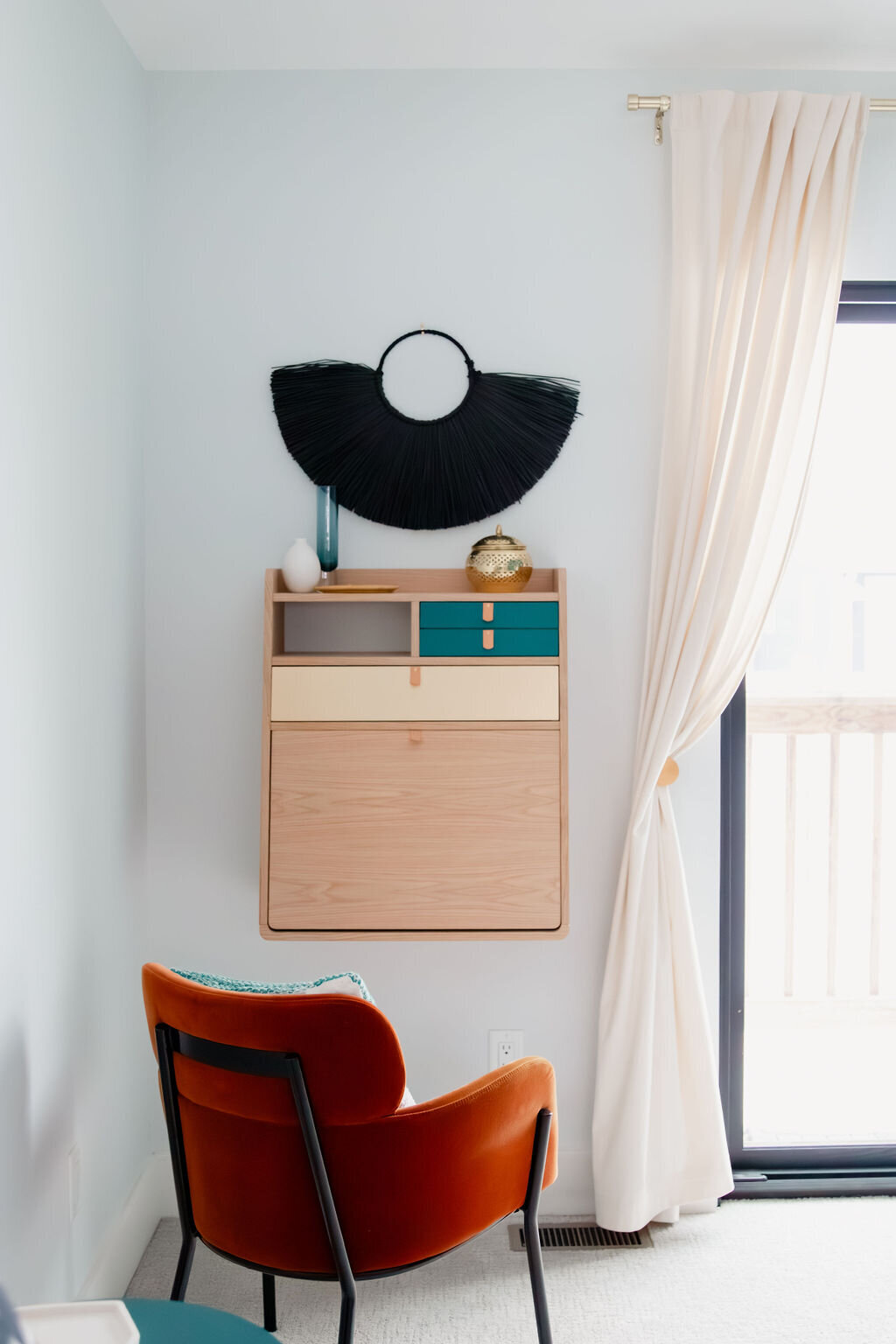 A modern bedroom corner with light blue walls features a light wood Gaston 60 Secretary Desk mounted on the wall. The desk has a drop-down writing surface and drawers with teal and cream fronts. Above the desk, a black fringed circular wall hanging is centered, flanked by a small light blue glass vase and a small gold decorative bowl. To the left, an orange velvet armchair with black metal legs and a patterned cushion sits on a light gray carpet. A sheer white curtain hangs to the right, partially covering a sliding glass door that leads to a balcony. Soft natural light fills the room.