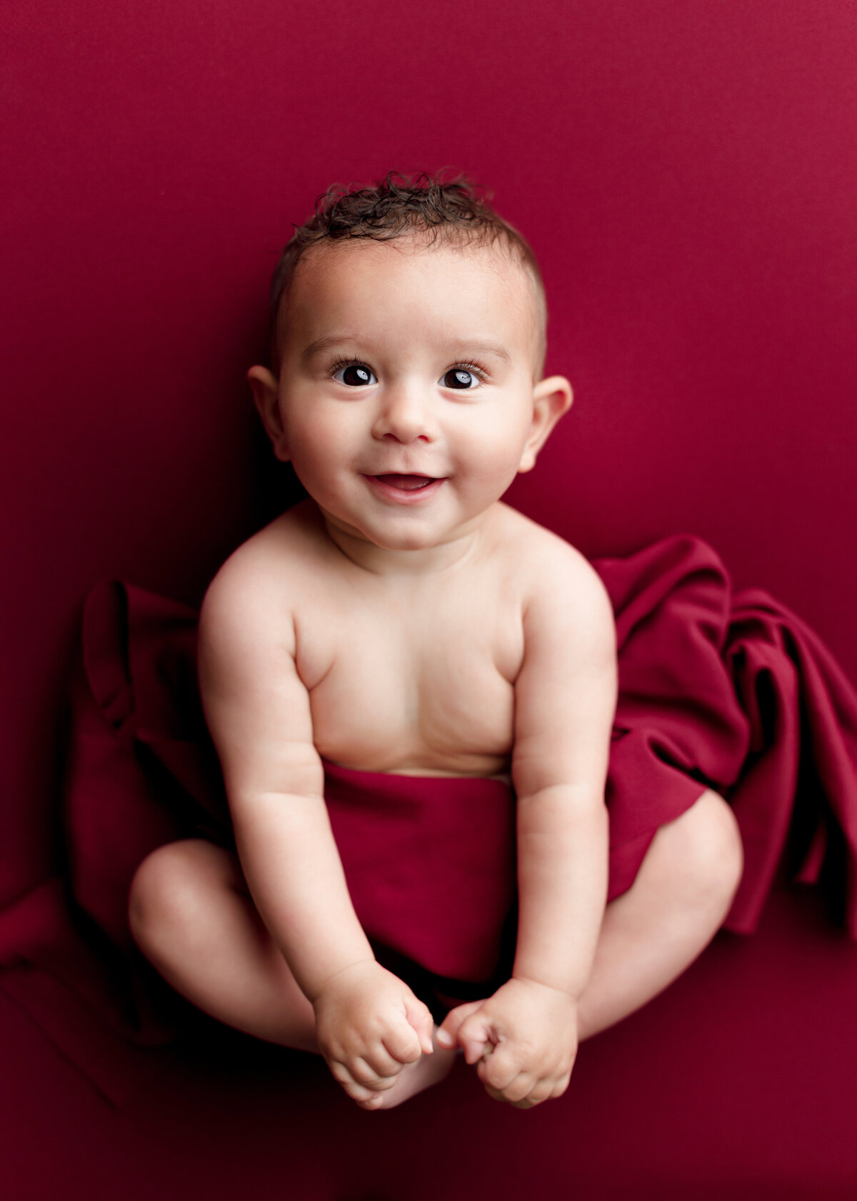 6-month milestone baby photos at top West Palm Beach and Wellington, FL photographer. Aerial image. Baby boy is bare and draped in red fabric against a red backdrop. Baby is holding his toes and smiling at the camera.