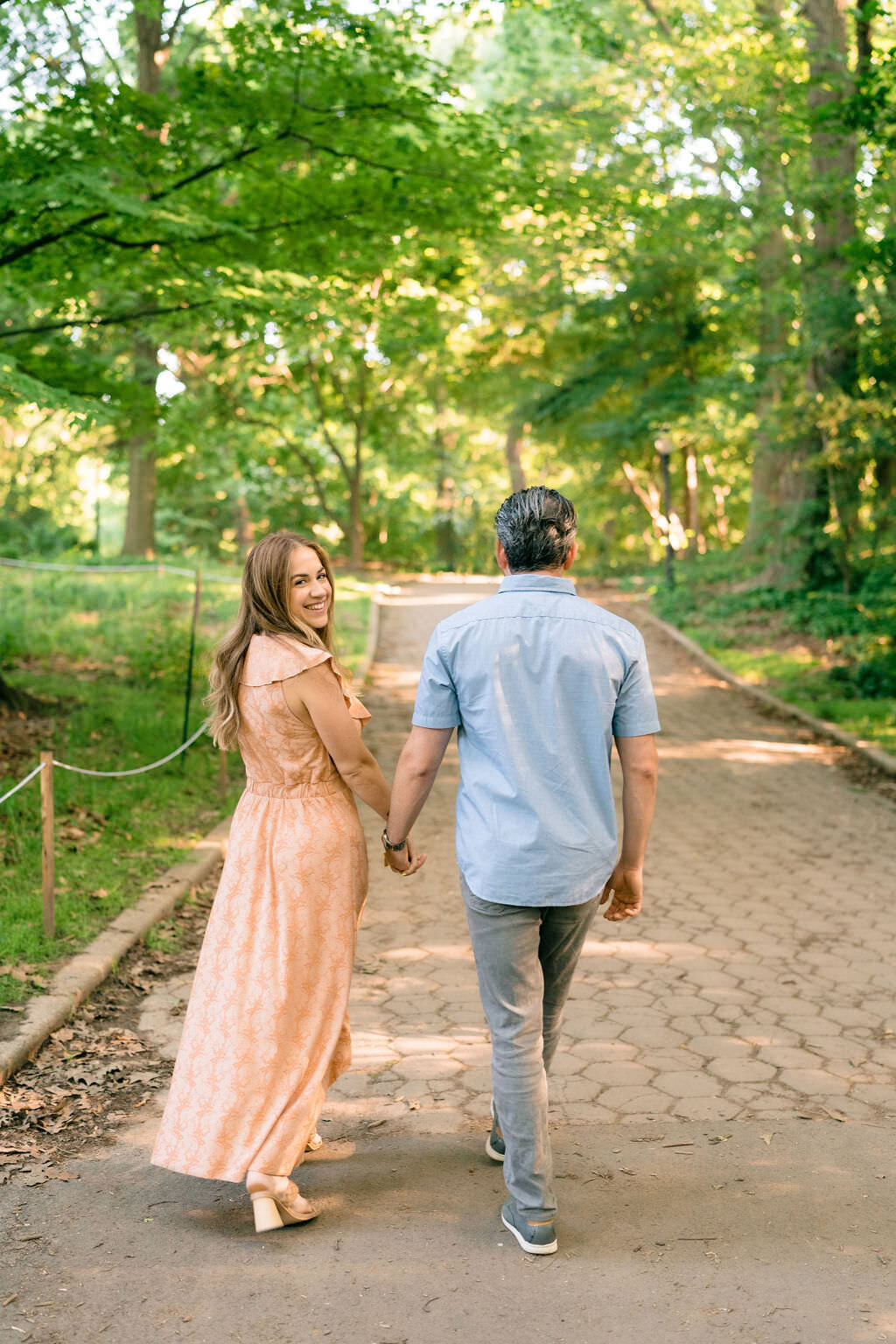 couple holding hands and walking along a path in the park as she looks back over her shoulder