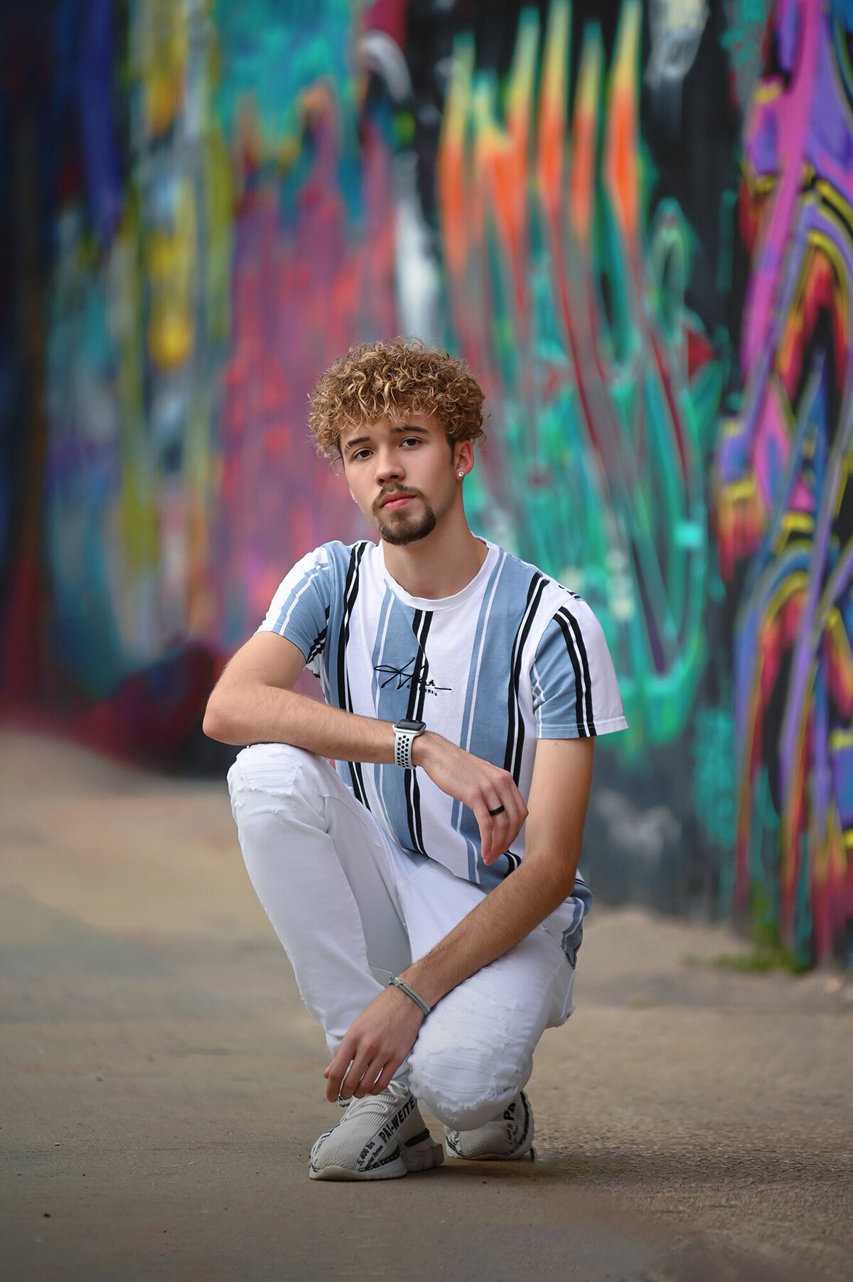 A high school grad kneels on the street in white pants and blue stripe shirt by some graffiti