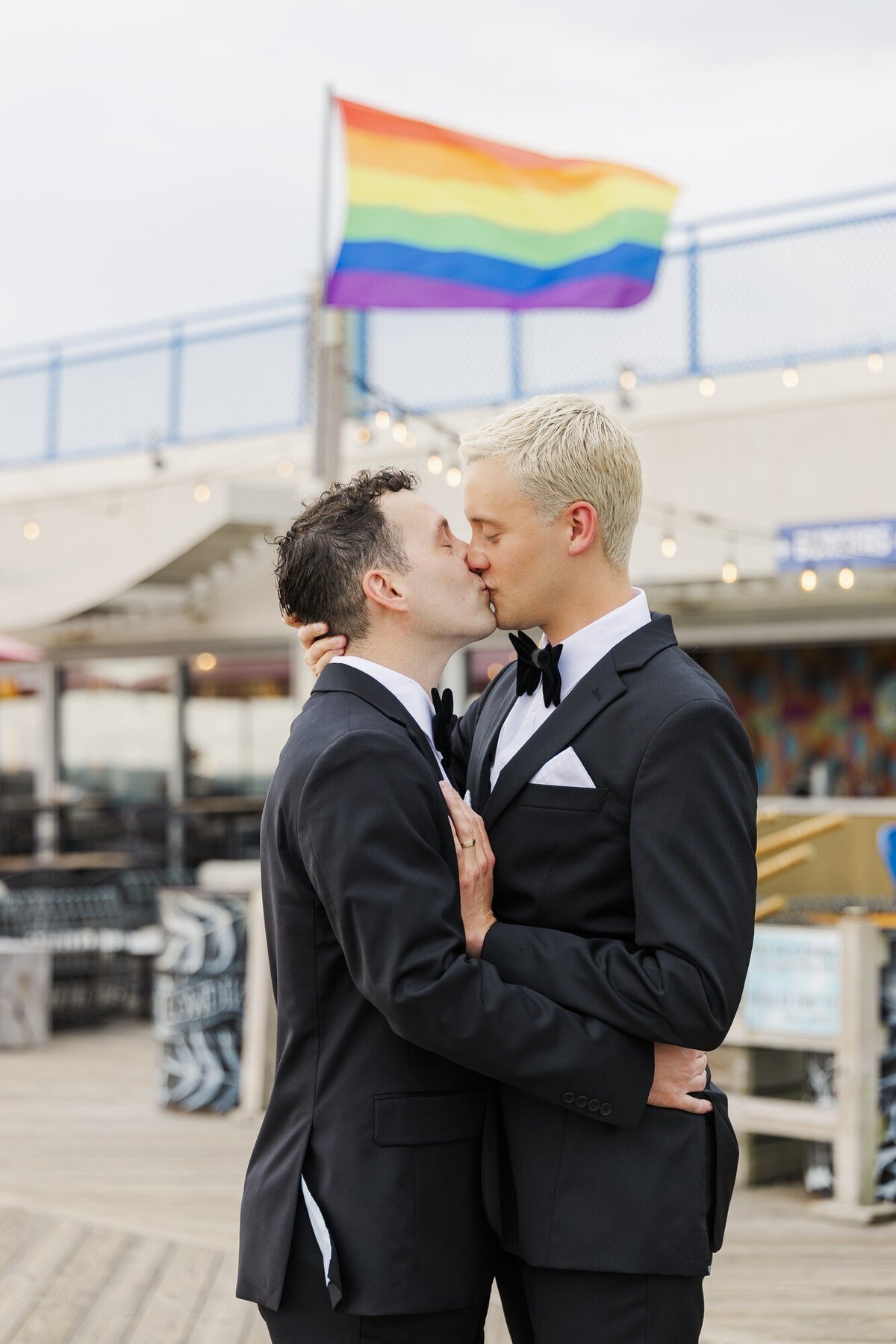 Two grooms in black tuxedos kissing in front of a rainbow pride flag on a waterfront boardwalk