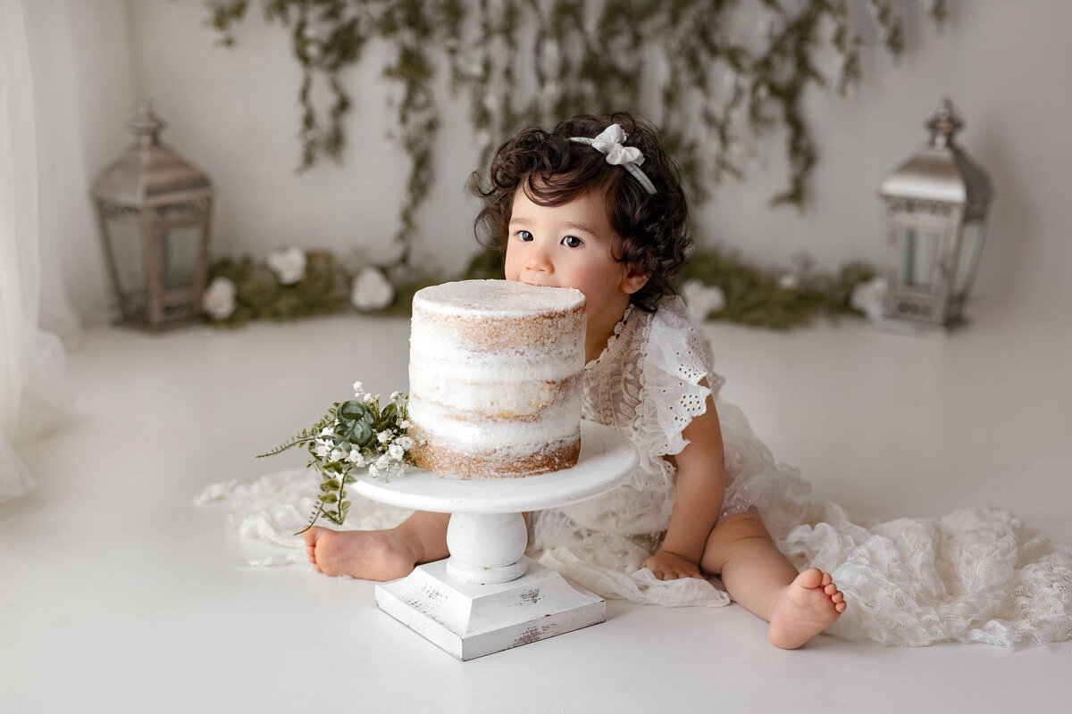 one year old girl eating her birthday cake in a white outfit and a white background with greenery in the background