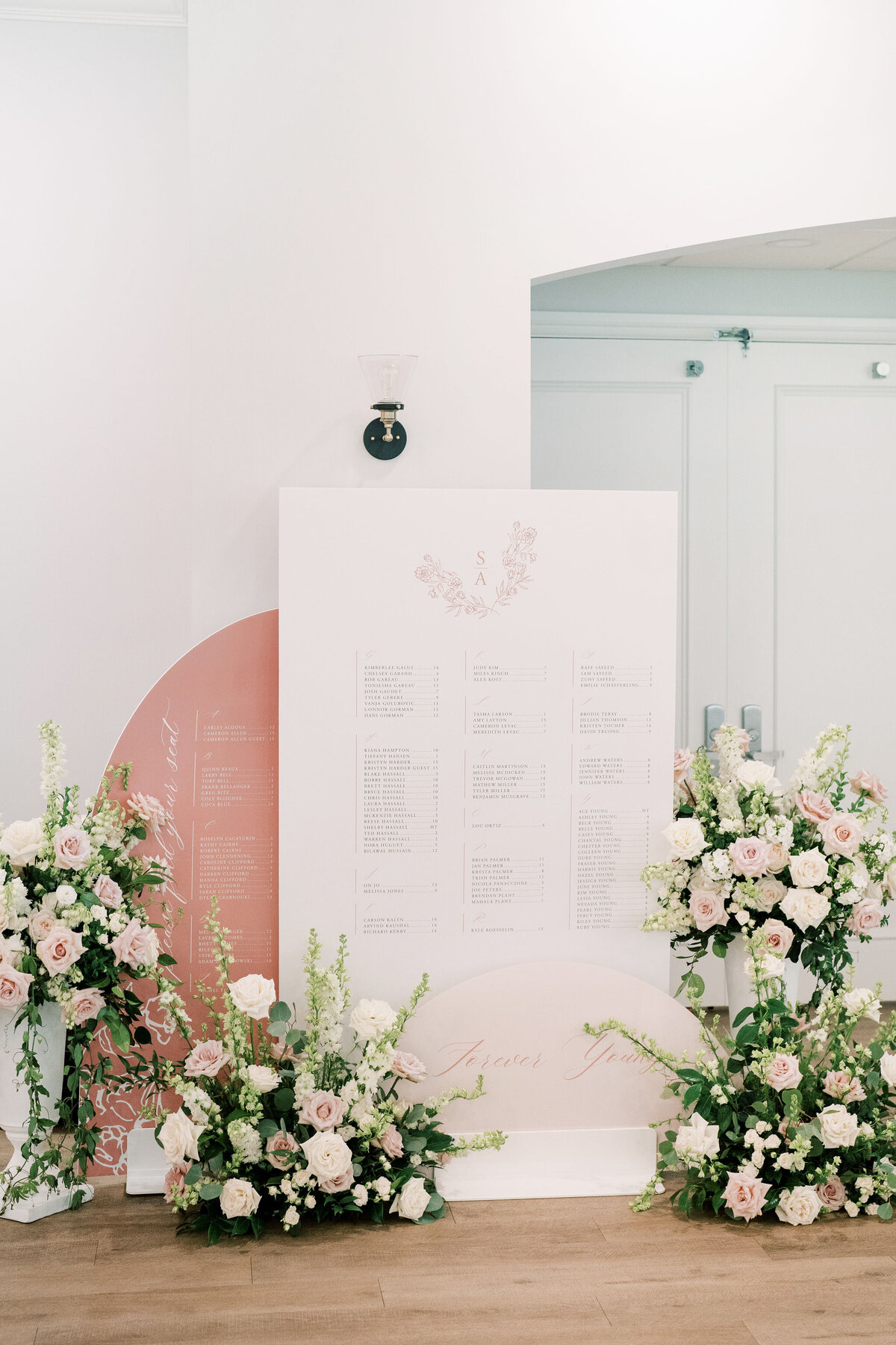 A large white seating chart adorned with floral arrangements including white roses and greenery stands against a white wall. The wedding design features multiple lists and is accented by a peach backdrop, perfect for an elegant wedding in Canada.
