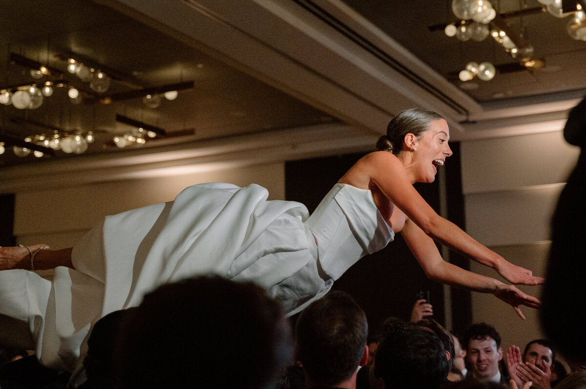 Bride being held aloft above wedding guests during the hora dance, arms outstretched, celebrating in Toronto.