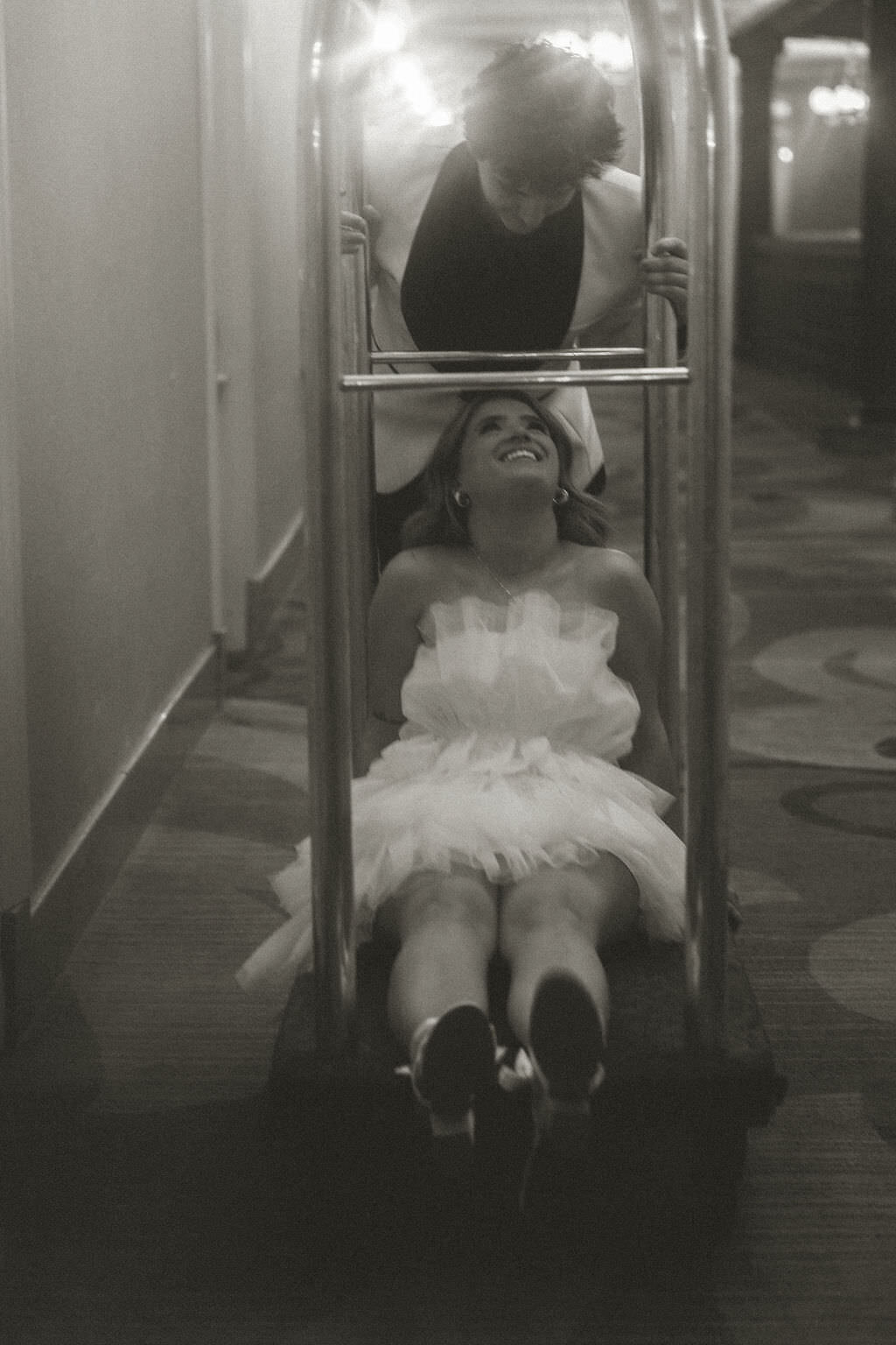 A person pushing their partner on a luggage cart.