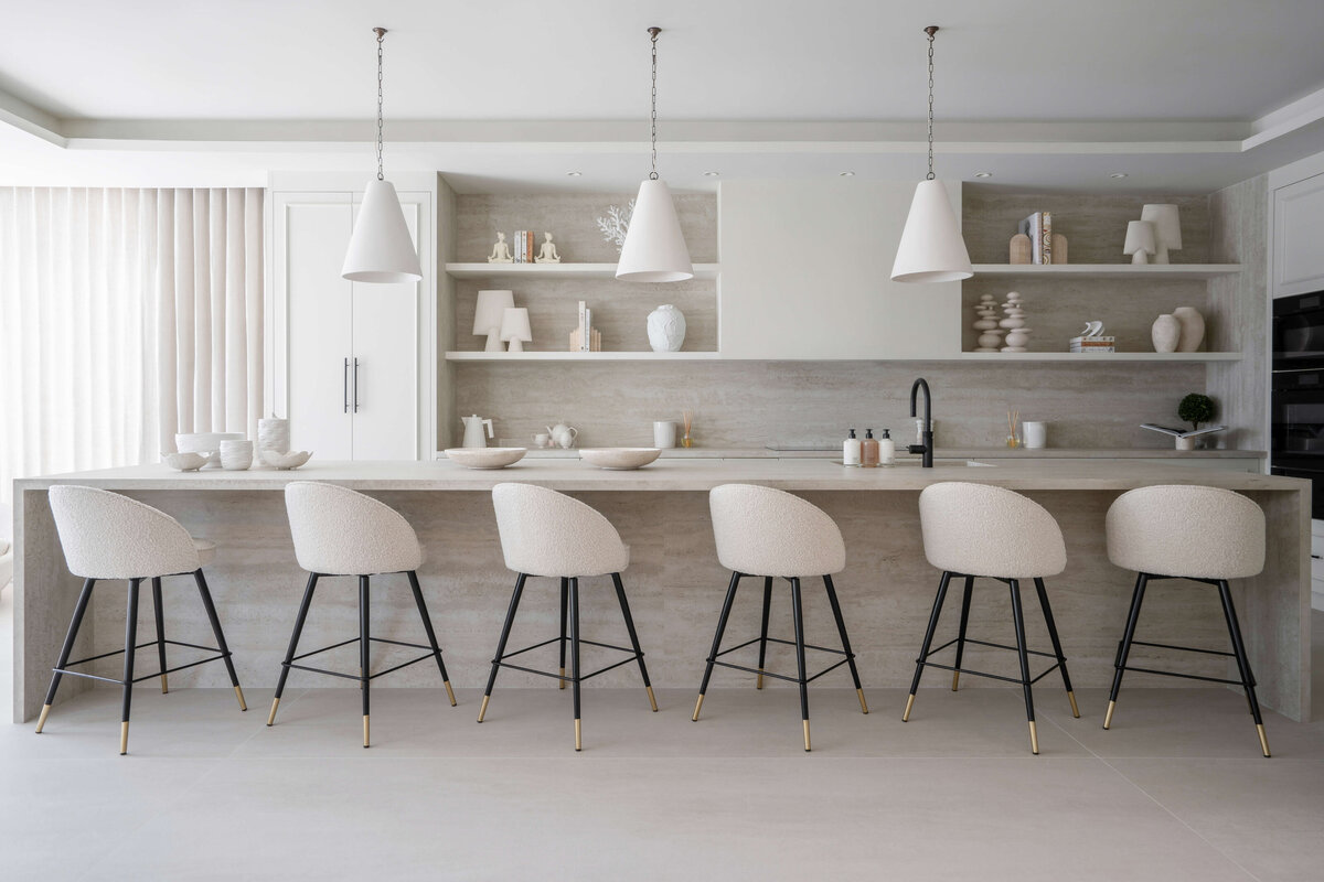 A large, open kitchen decorated with a bright, minimal aesthetic. Along a long bar sit six white rounded barstools with black legs. Above are three white pendant lights. Behind, there  is built-in white shelving with minimal white chic decor.