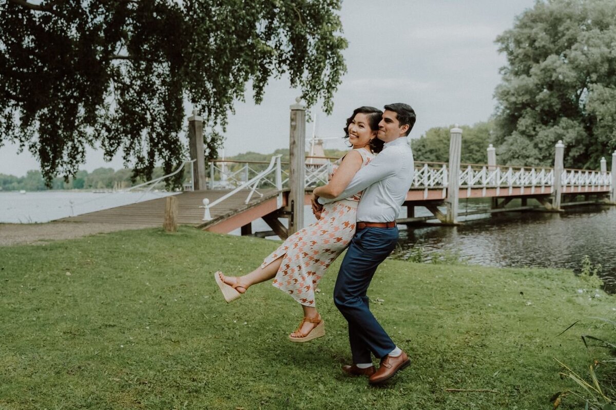 A playful and romantic candid photo of a couple in a park. The man, in a white shirt and blue pants, joyfully lifts the woman off the ground by her waist. She wears a floral, patterned dress and is laughing with her legs playfully bent. In the background, there is a wooden bridge over a body of water and lush green trees.