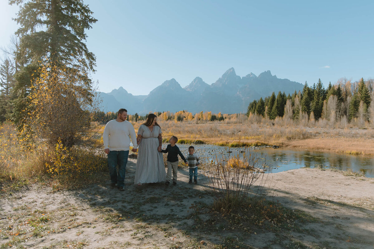 A small family consisting of a mom, dad, and 2 young boys all hold hands as they walk towards the camera looking at one another. Behind them are the Grand Tetons.