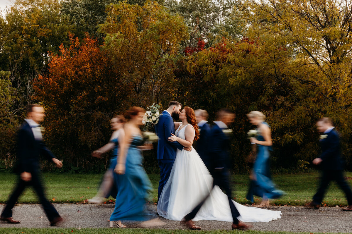 A Bride and groom kiss while the wedding party walks around them.