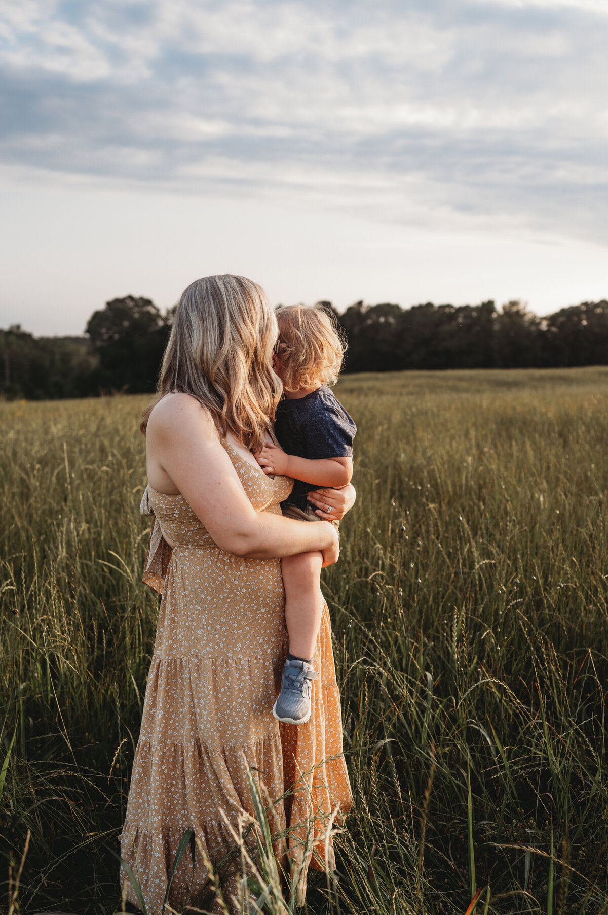 Mother holds her little boy in a large open field while gazing into the sunset