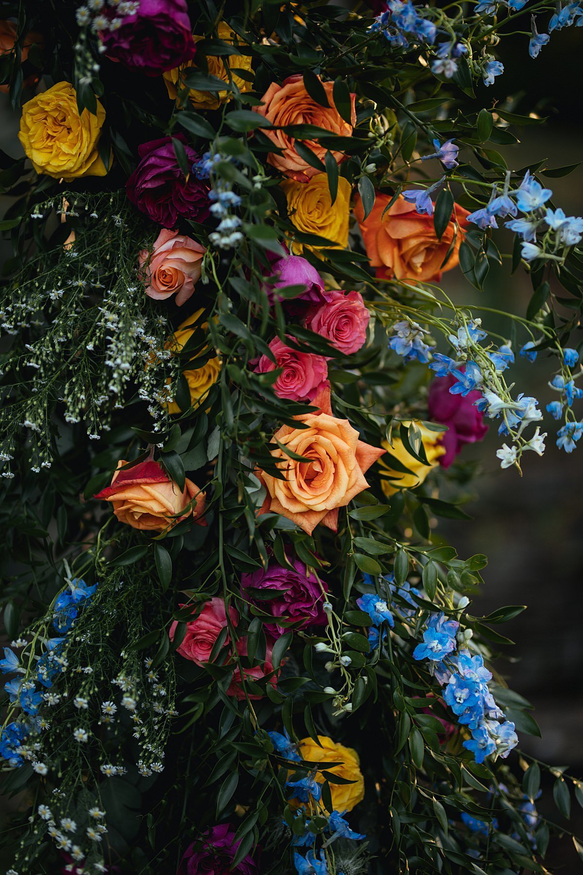 A detail photo of an elaborate and lush floral arbor covered in heavy greenery with blue, orange, peach, hot pink , burgundy and purple flowers