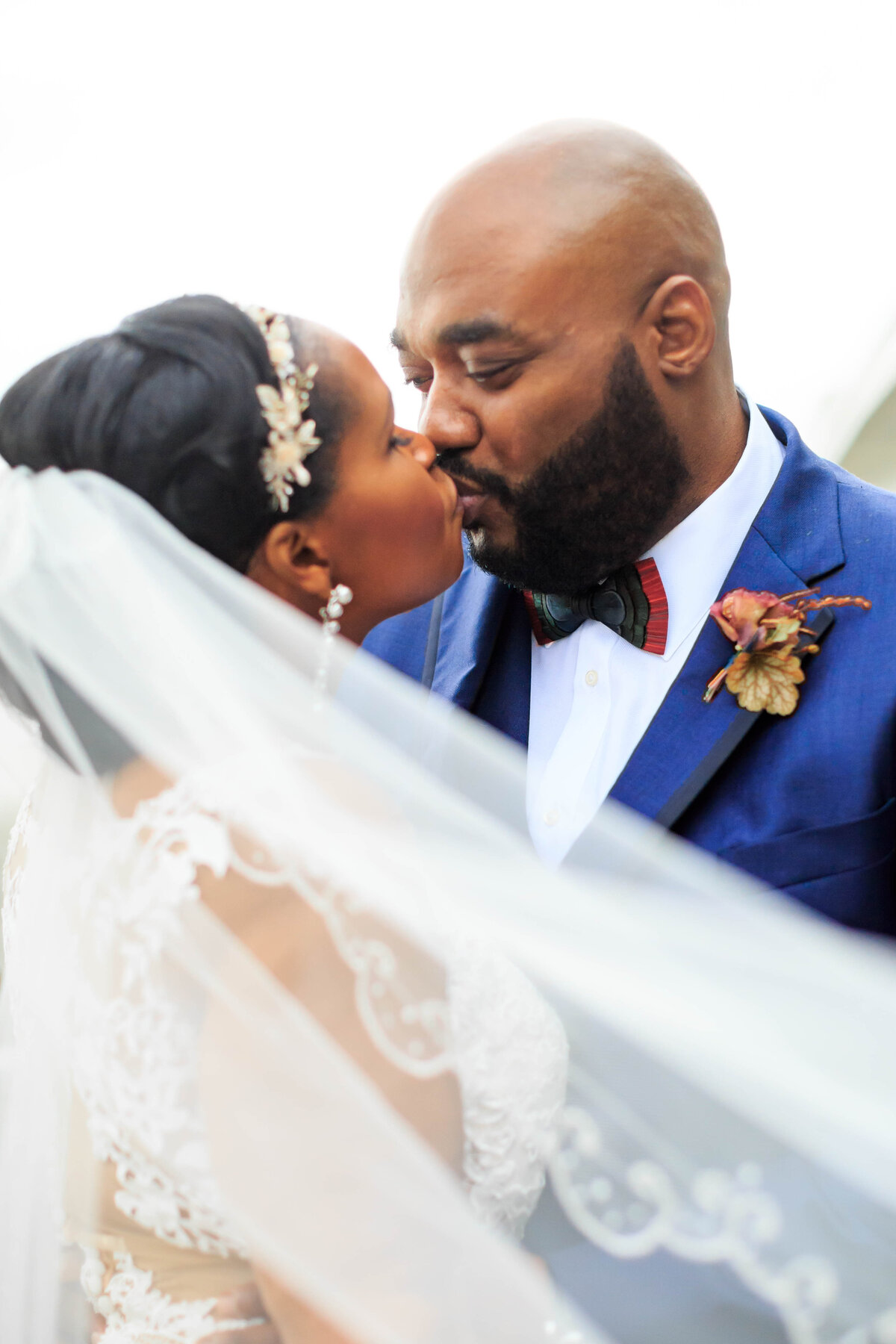 Couple kiss sweetly in a closeup portrait, featuring a lace veil and his blue suit.