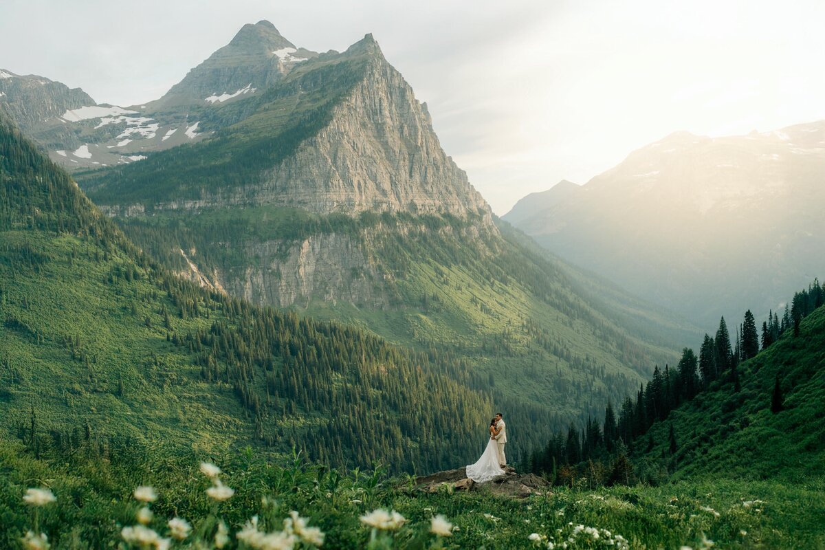 Romantic wedding couple embracing on a mountain overlook surrounded by wildflowers in Glacier National Park Montana with dramatic alpine peaks and golden sunset light in the background