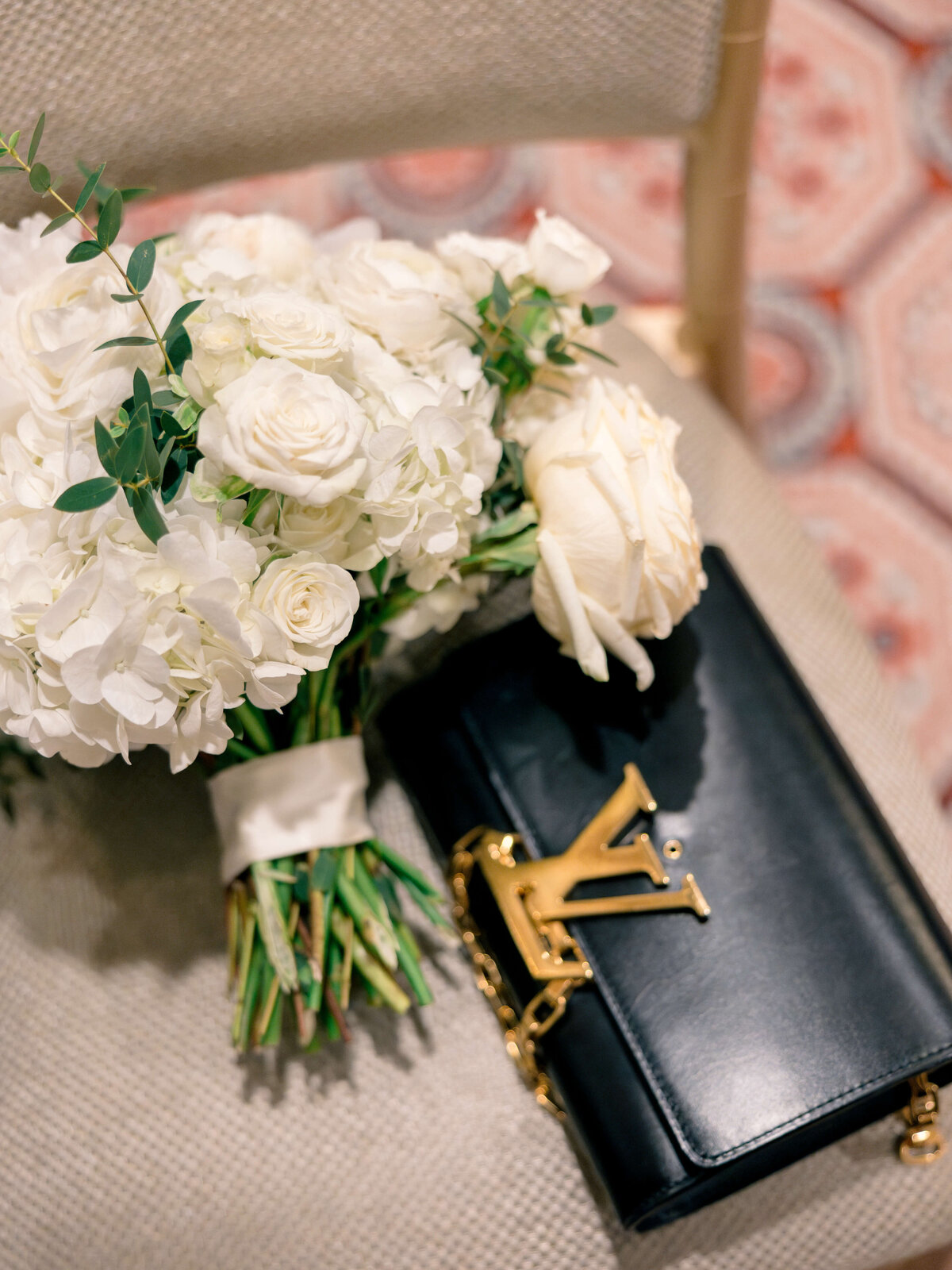 A bouquet of white roses and hydrangeas with greenery lies on a textured chair next to a black designer handbag featuring a gold LV emblem. The background shows a blurred, patterned rug.