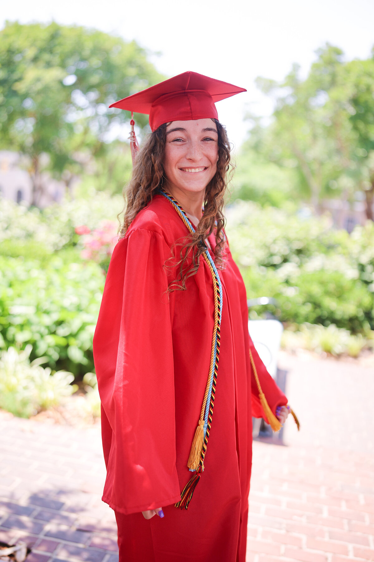 female high school graduate wearing red cap and gown in park with flower