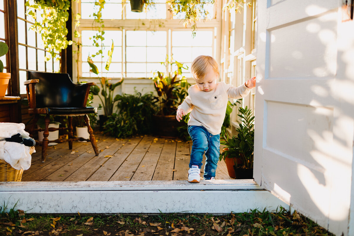 A baby boy is walking in a wooden house with white door. He is dressed with a jean and white coat. Behind him are plants and a blue chair