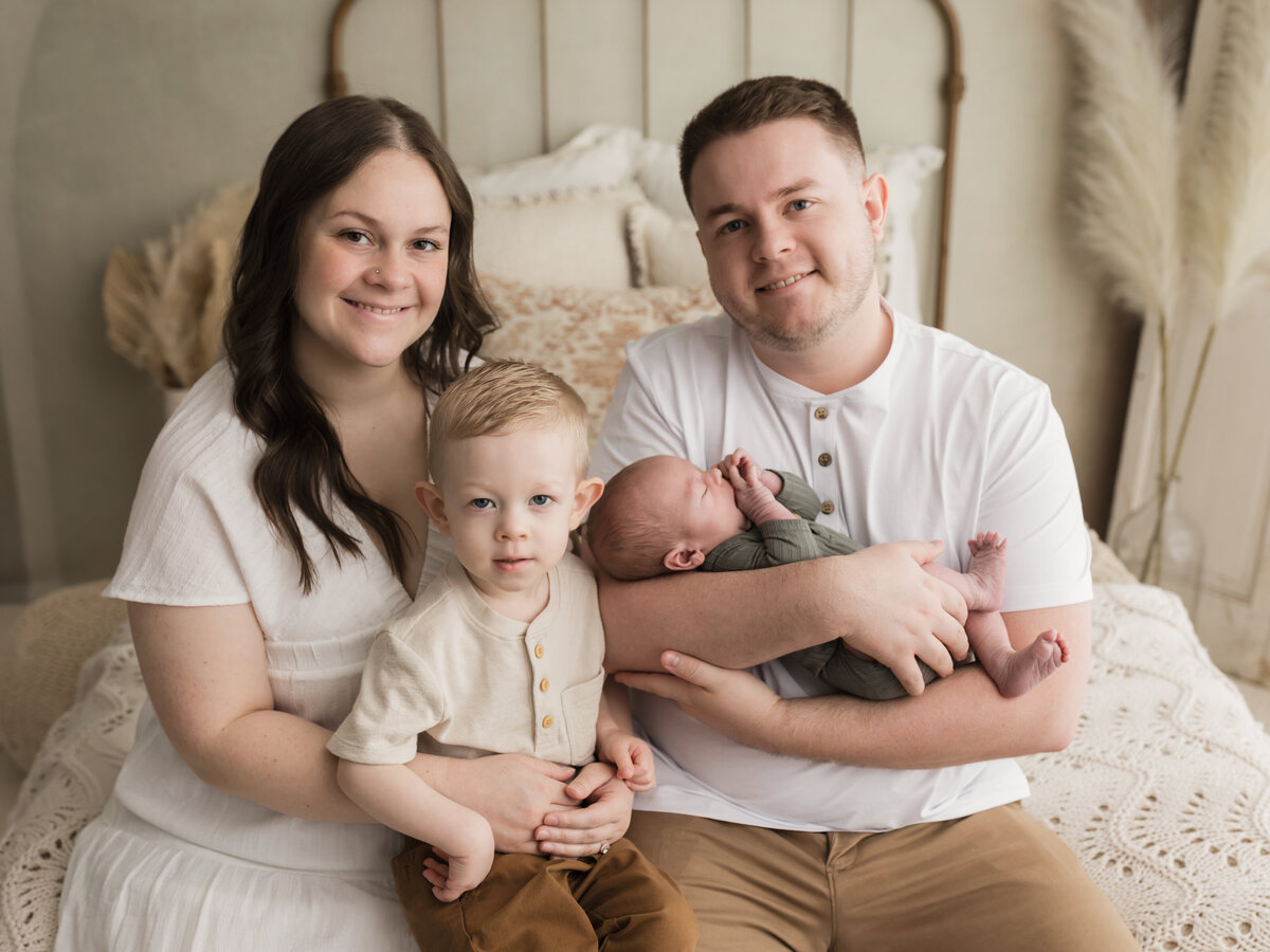 parents sitting on bed holding two year old son and newborn baby boy for studio portraits cleveland newborn photographer
