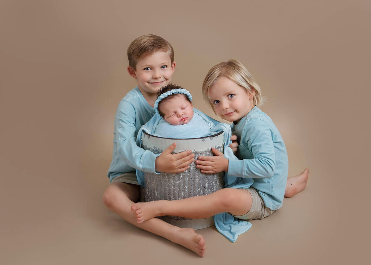 Two older brothers with their newborn baby sister in bucket all wearing blue taken by Los Angeles newborn photographer