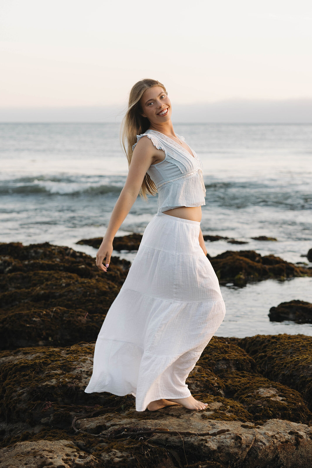 Senior girl in a white skirt on ocean rocks at sunset