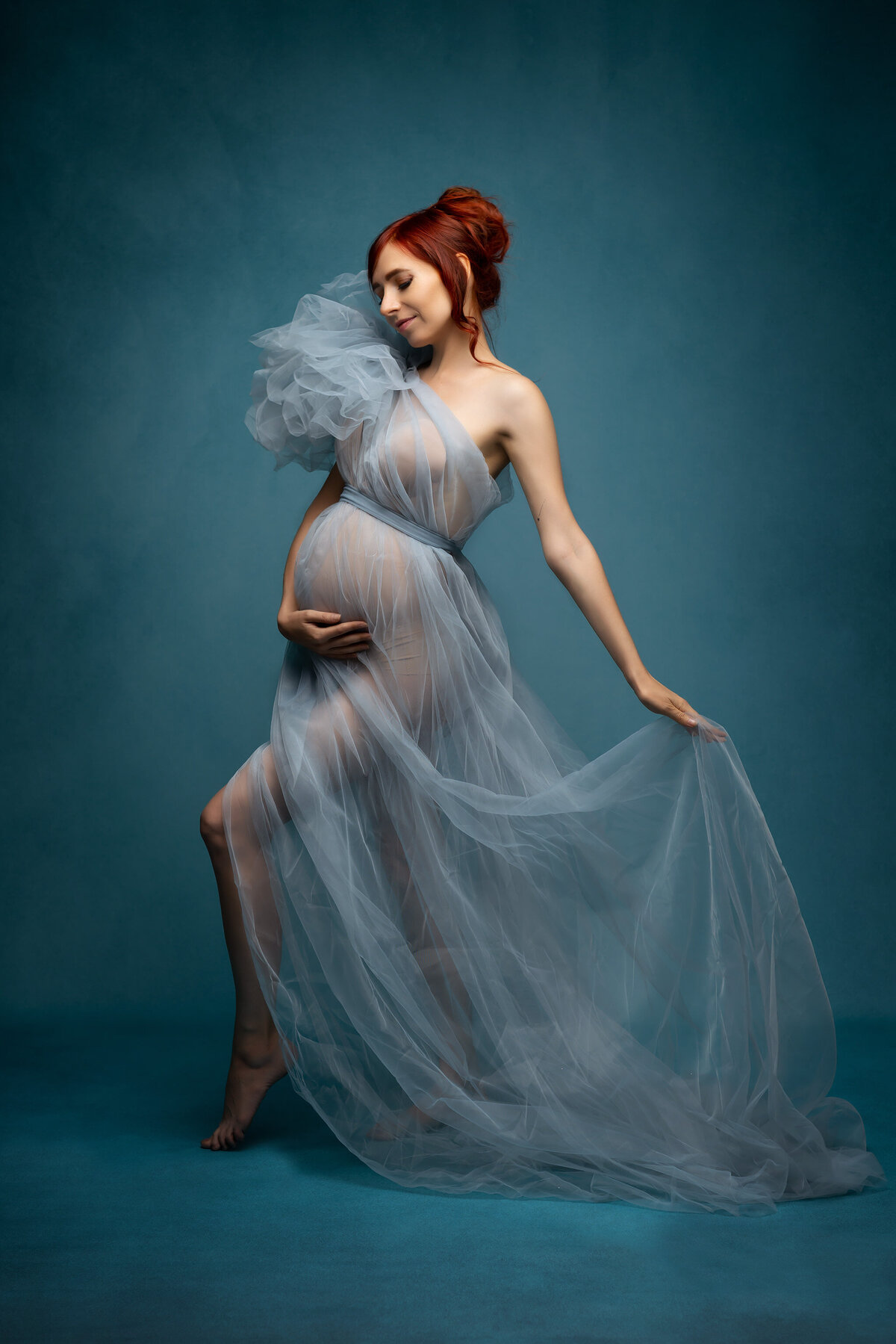 Stunning monochromatic  teal maternity photograph of a pregnant woman wearing a teal tulle gown with an asymmetrical sleeve. She has striking red hair.