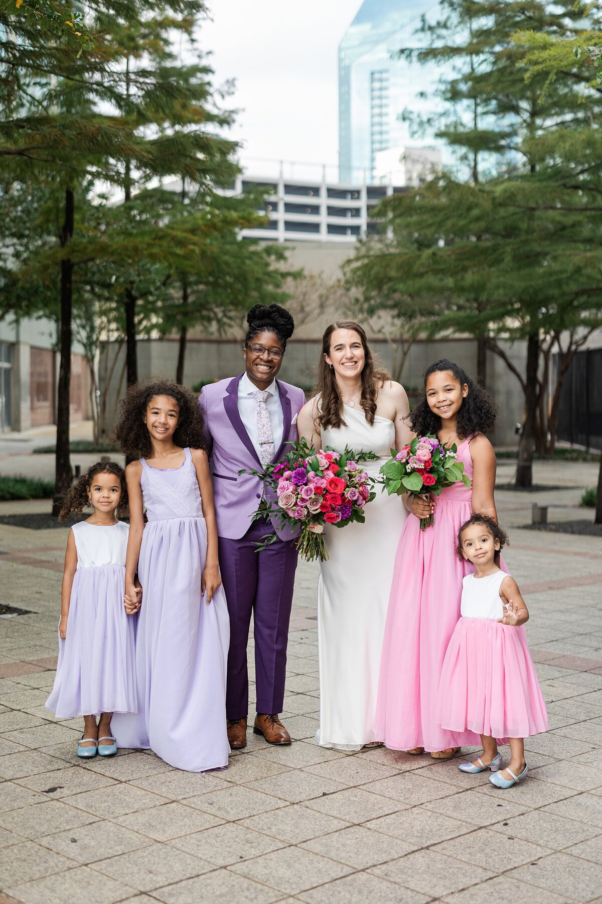 A portrait of two brides and the child attendants from their wedding party after their wedding ceremony at The Westin Dallas Downtown in Dallas, Texas. The bride on the left is wearing a purple suit with a floral tie and her two child attendants are wearing lavender dresses. The bride on the right is wearing a sleeveless, elegant, white dress with a large bouquet, and her child attendants are wearing pink dresses.