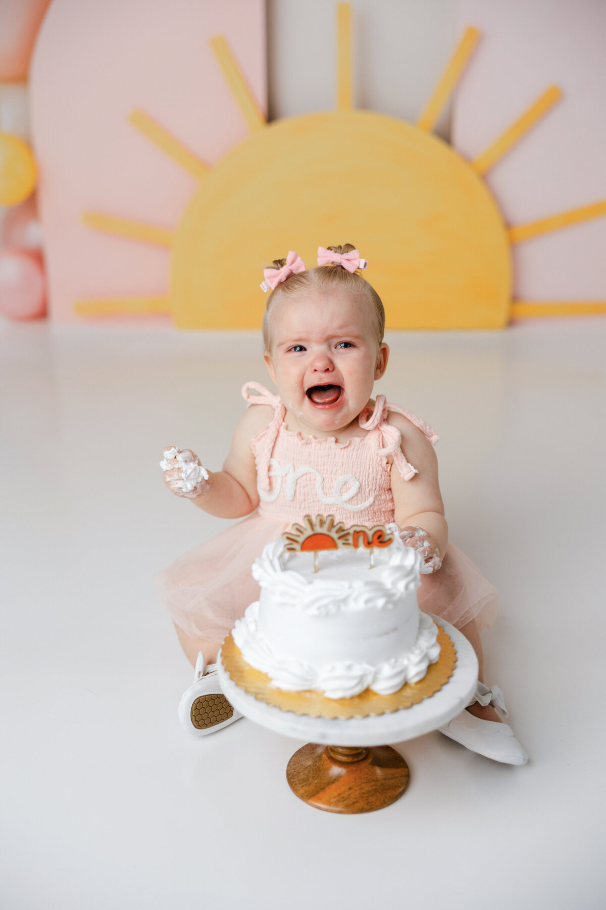 Baby in peach “one” dress crying during cake smash session with sunrise and balloon backdrop. Photograph by Yucaipa cake smash photographer Kaitlyn Dawn Photography.