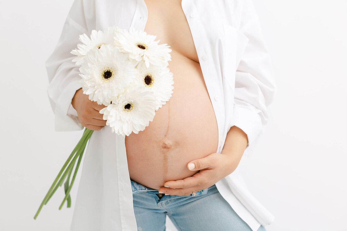 Close-up image of a baby bump with Mama holding white flowers next to them. Mama is wearing a white buttoned down shirt that is open and showing off her baby bump.