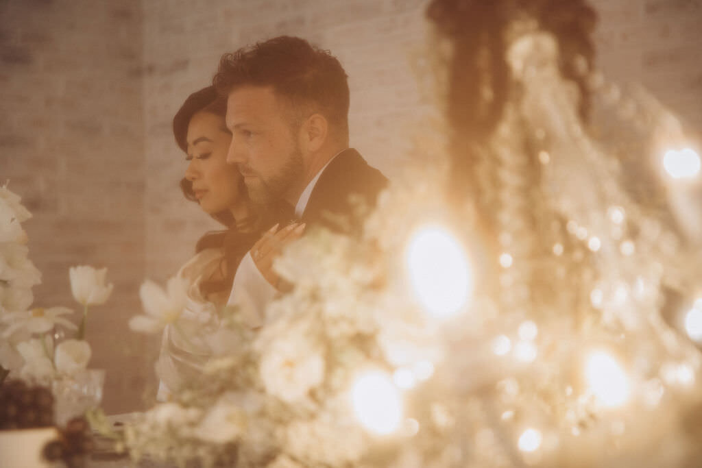 A copule hugging from behind with a chandelier in front of them.