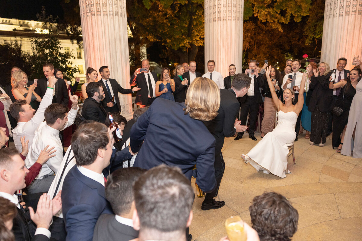 A bride sits smiling on a chair while a groom kneels in front of her, surrounded by cheering guests at an outdoor wedding reception. The setting includes elegant columns and trees in the background.