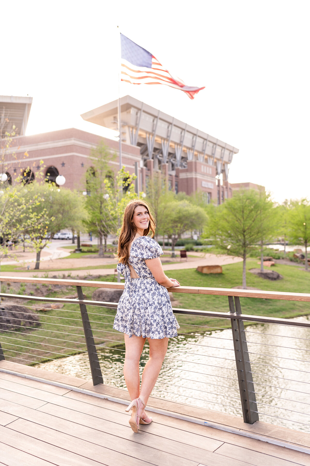 Texas A&M senior girl holding on bridge railing and looking over shoulder in blue dress in Aggie Park with Kyle Field in background