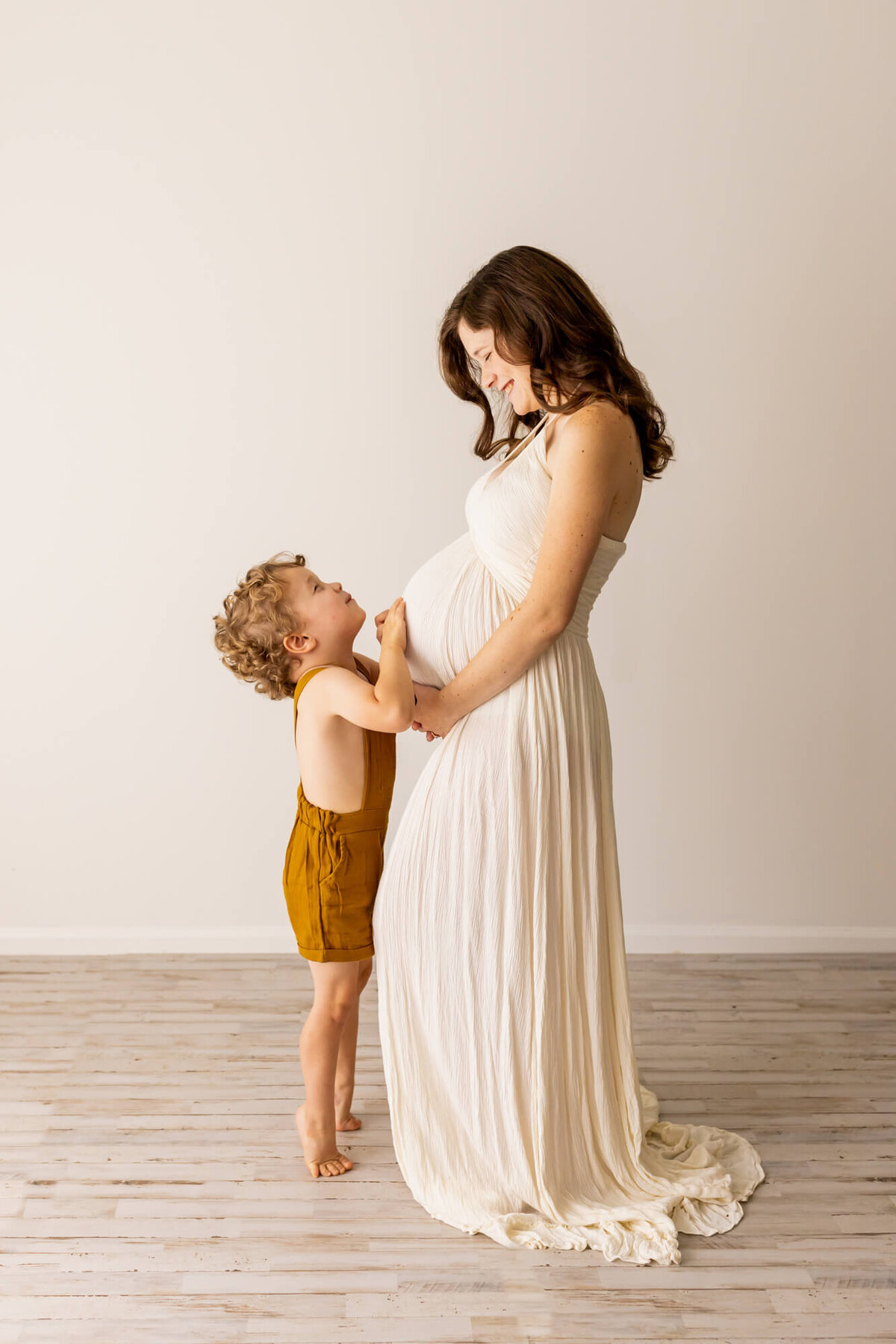 Mom to be in a white boho dress looking at her toddler in a mustard jumpsuit