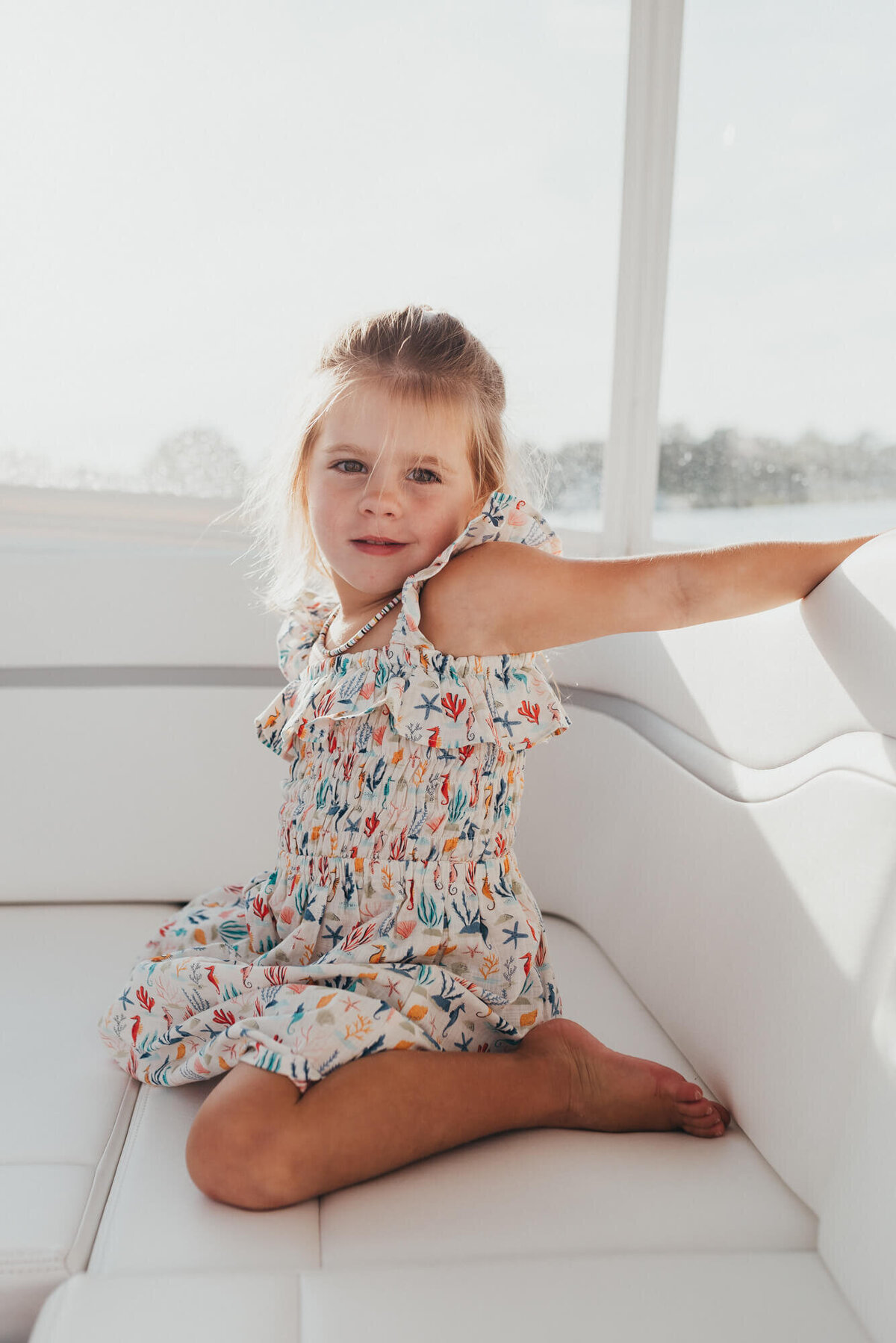 A little girl sits on her family's boat, she is wearing a cute ocean themed dress and looking at the camera