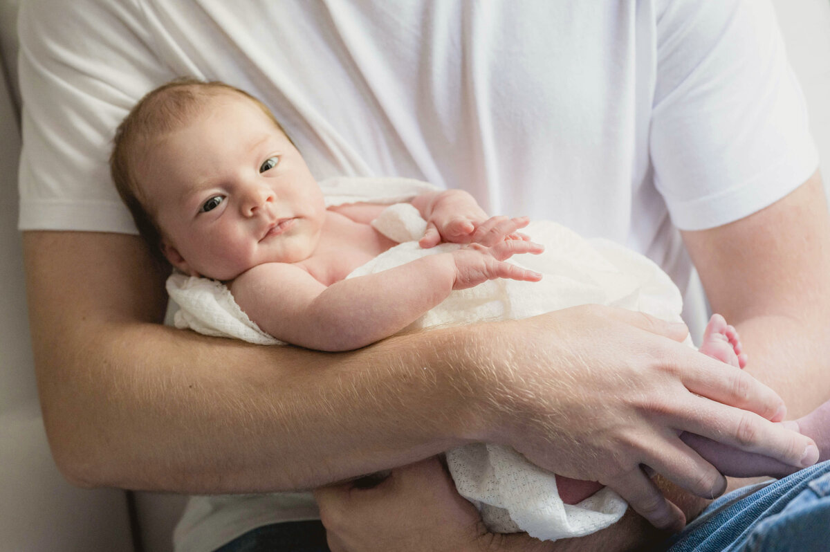 A week old baby wrapped in a white blanket being held in  her father's arms and looking at the camera,.