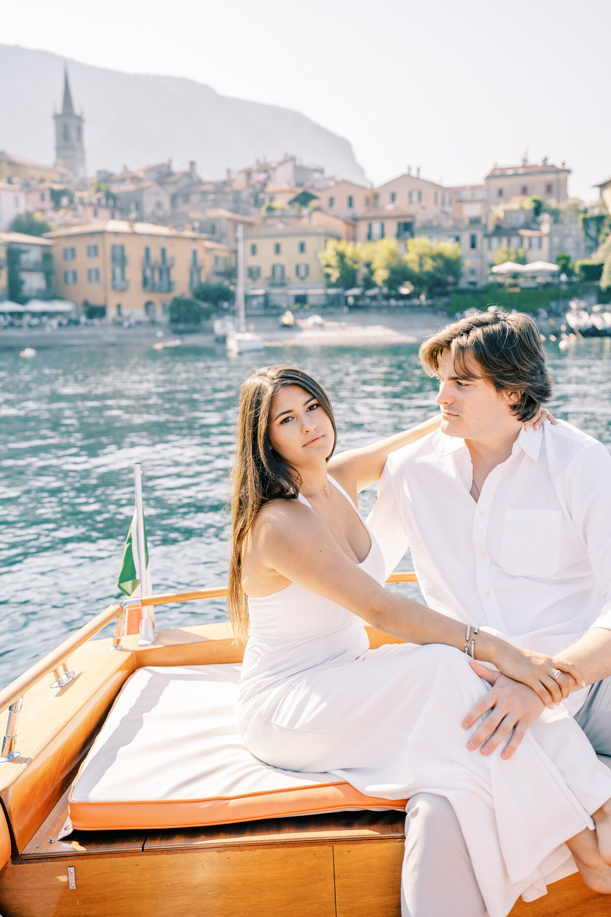 Couple in all white in a boat with the town of Varenna in the background at their Italy engagement session, photographed by Italy wedding photographer.
