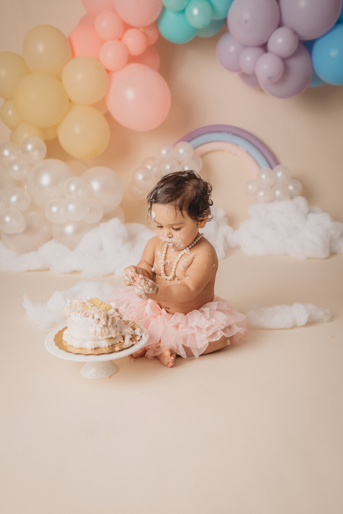 one year old baby girl at atlanta ga cake smash portrait studio wearing a pink tutu and smashing and eating a cake  with pastel rainbow balloon garland behind her