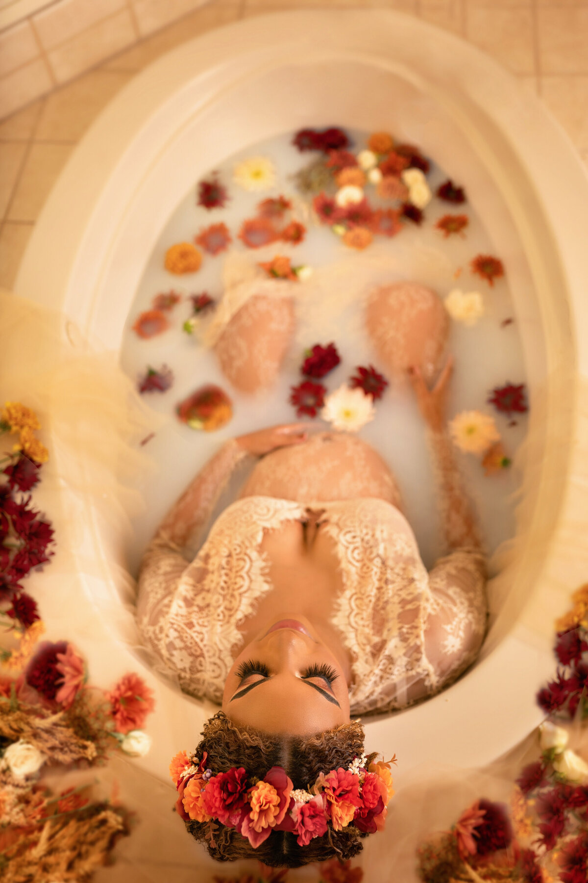 Black woman with a floral crown wearing a beige lace dress that shows off her pregnant belly.  She is leaning back against the bathtub and has her eyes closed.  Her hair is pulled back and she has long eyelashes.  There are yellow, red, and white flowers floating in the milk water.
