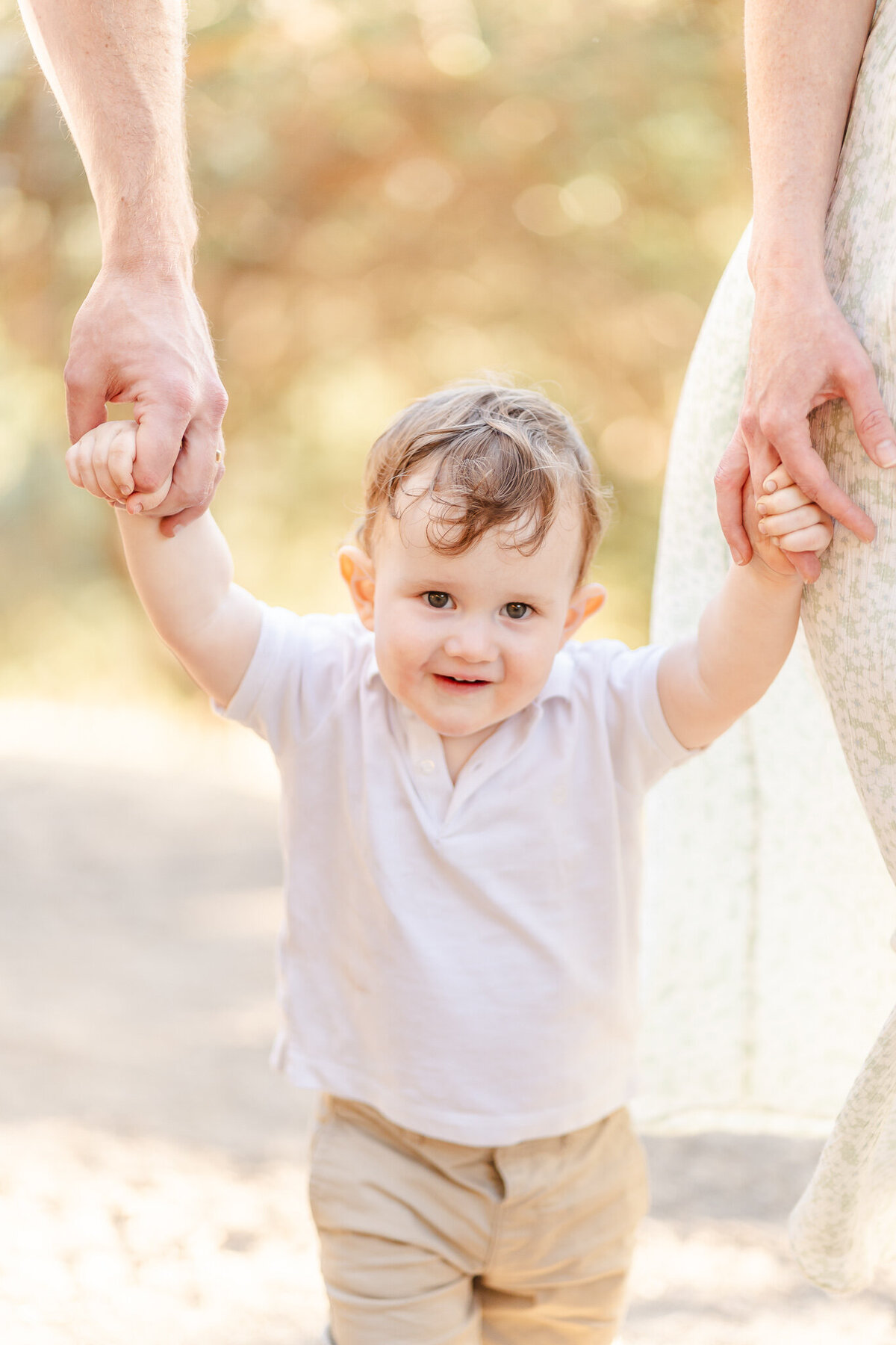 closeup of toddler boy in white shirt holding his parents' hands while walking and looking at the camera