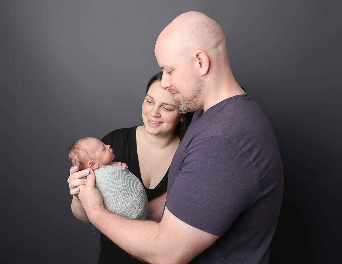 A loving mom and dad hold their newborn son, gazing down at him in a cozy photography studio in Rochester, NY, capturing the warmth of family love.