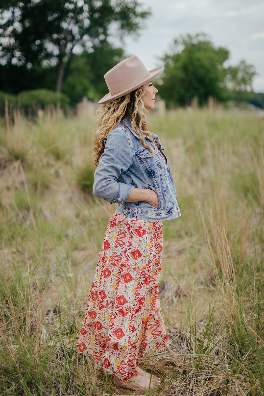 woman in tall grasses is wearing a red floral dress and jean jacket, and a fedora. She is in profile looking into the distance as the breeze blows her dress and hair.
