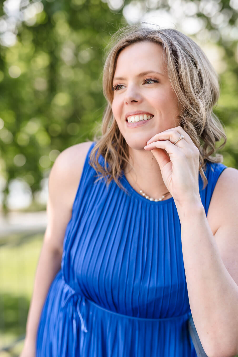 Smiling women’s health doctor in pleated blue dress with pearl necklace outdoors in Kelowna's City Park.