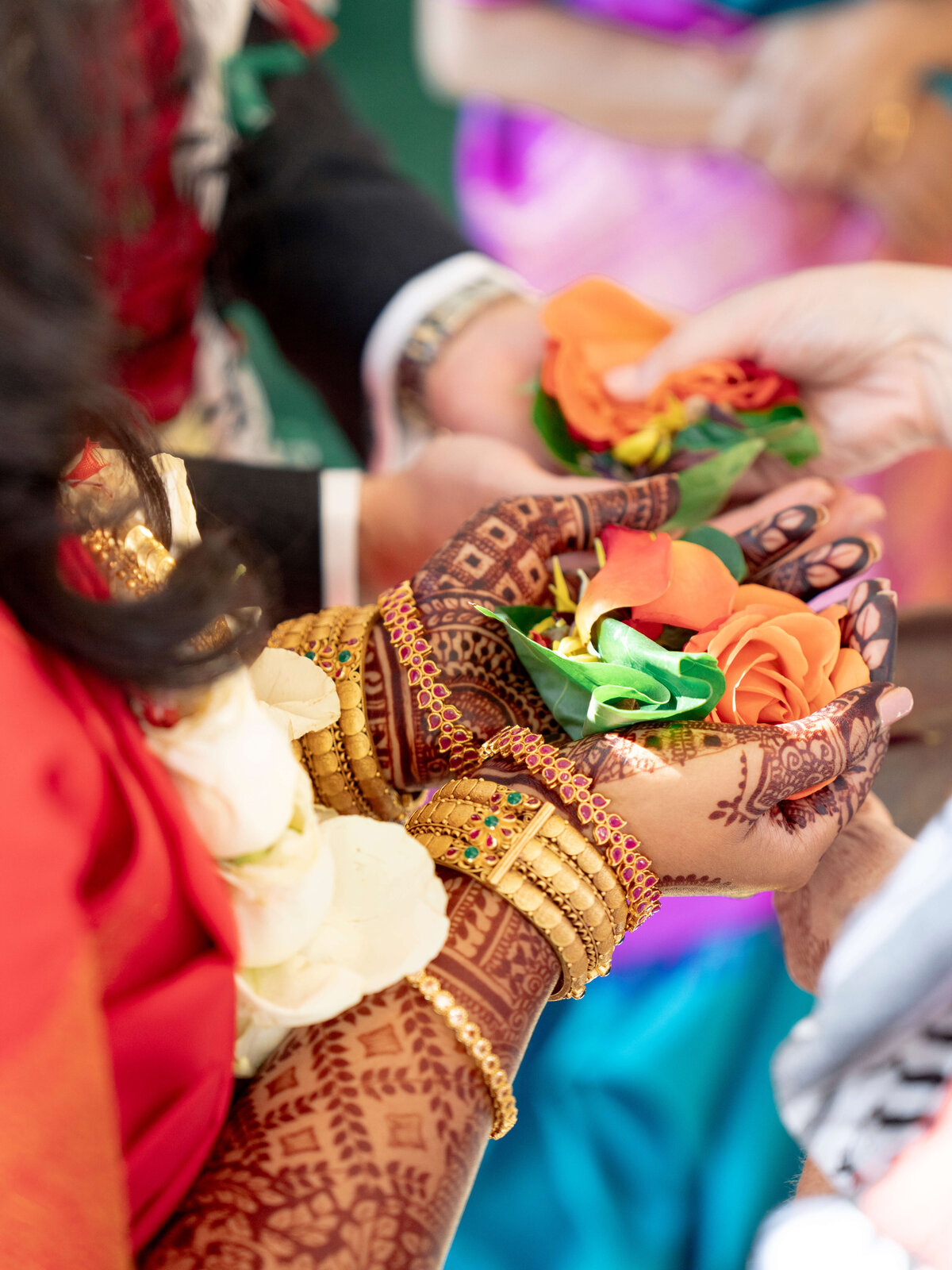 A close-up of hands adorned with intricate henna designs and gold bangles holding orange and yellow flower decorations, set against a background of blurred colorful fabrics.